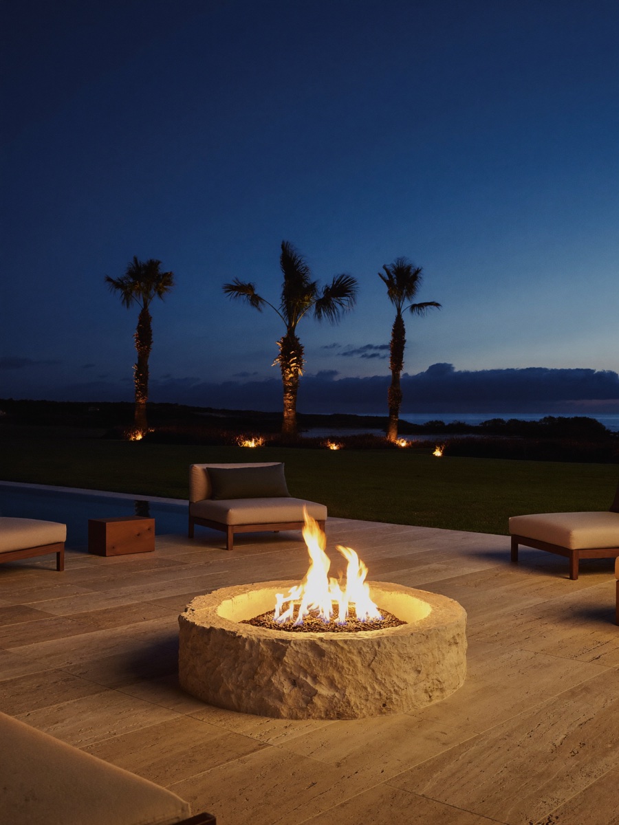 A custom natural-stone fire pit on a travertine paver patio glowing at dusk.
